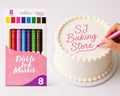 An 8-pack of edible ink markers shown next to a white frosted cake on a white surface, with a hand using a pink marker to write 'SJ Baking Store' on the cake.